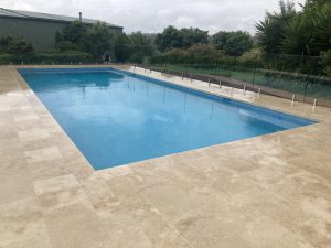 Blue swimming pool with glass fencing and ivory paving
